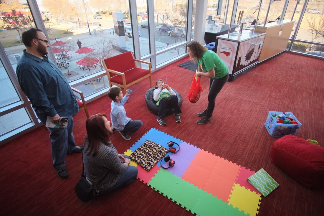 A mother and father standing up and looking down at their two sons sitting on bean bags at the Durham Performing Arts Center. There are also headphones and a foam mat on the ground